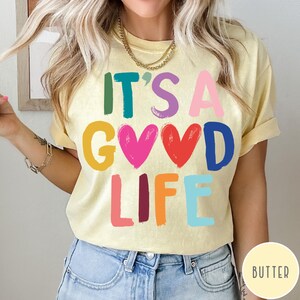 K&ouml;nnte beinhalten: Ein blassgelbes T-Shirt mit dem mehrfarbigen Schriftzug IT'S A GOOD LIFE, bei dem zwei Herzen die beiden O im Wort GOOD ersetzen. Die Buchstaben sind in verschiedenen Farben gedruckt, darunter Petrol, Lila, Pink, Gelb, Rot, Blau und Orange.