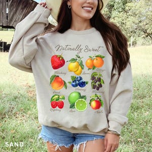 May include: A beige sweatshirt with the text "Naturally Sweet" printed on the front. The sweatshirt features a graphic of various fruits and berries, including a strawberry, peach, lemon, orange, blueberry, blackberry, raspberry, lime, and apple.