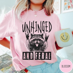 May include: Pink t-shirt with a black and white illustration of a raccoon with the text "Unhinged and Feral" printed on it.