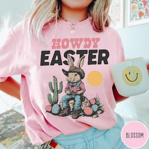 Puede incluir: Sudadera rosa claro con las palabras "HOWDY EASTER" en rosa y negro. Un conejo de dibujos animados con sombrero de vaquero y sentado en una roca aparece debajo del texto. El conejo est&aacute; rodeado de huevos de Pascua y un cactus. Una taza blanca con una carita sonriente se sostiene en la mano.