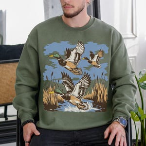 May include: Olive green sweatshirt featuring a graphic of three ducks in flight over water and reeds. The ducks are depicted with brown, white, and green feathers, with yellow beaks. The background includes blue sky and brown reeds.