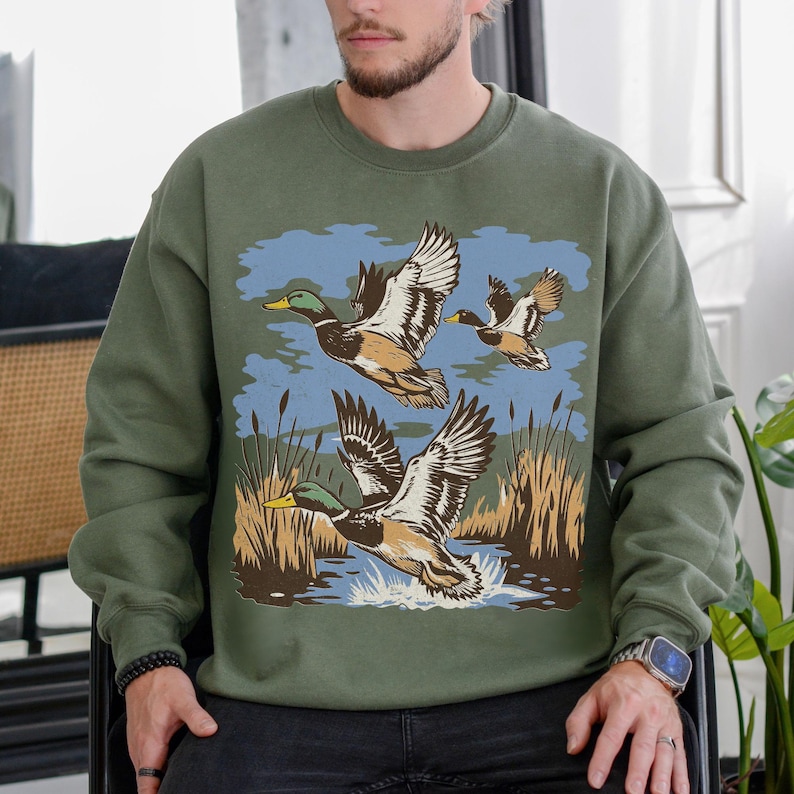 May include: Olive green sweatshirt featuring a graphic of three ducks in flight over water and reeds. The ducks are depicted with brown, white, and green feathers, with yellow beaks. The background includes blue sky and brown reeds.