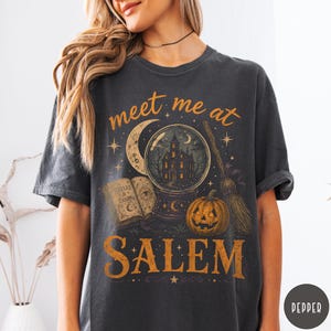 May include: A charcoal gray short-sleeve t-shirt featuring a graphic print with the text "meet me at SALEM". The design includes a crystal ball with a haunted house, a crescent moon, an open book titled "SPELLS & CHARMS", a pumpkin, and a broom.