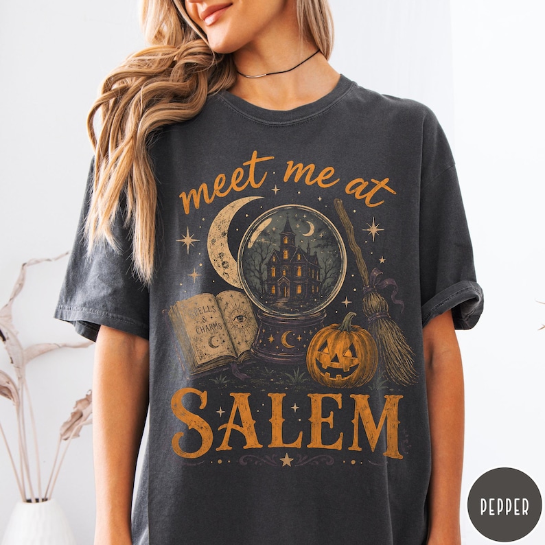 May include: A charcoal gray short-sleeve t-shirt featuring a graphic print with the text "meet me at SALEM". The design includes a crystal ball with a haunted house, a crescent moon, an open book titled "SPELLS & CHARMS", a pumpkin, and a broom.