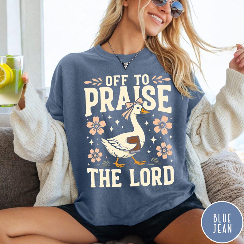 Puede incluir: Camiseta azul vaquera con el texto "OFF TO PRAISE THE LORD". El dise&ntilde;o presenta un ganso con un lazo y un libro, rodeado de flores. La camiseta tiene un corte holgado.
