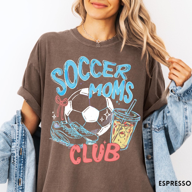 May include: Espresso-colored t-shirt with a graphic that reads "Soccer Moms Club" in blue and pink. The design includes a soccer ball, cleats, and a beverage. The shirt is a casual style.
