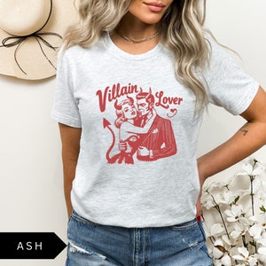 May include: A heather grey t-shirt with a red graphic print of a couple. The man has horns and a suit, and the woman is wearing a dress. The text "Villain Lover" is printed above the couple.