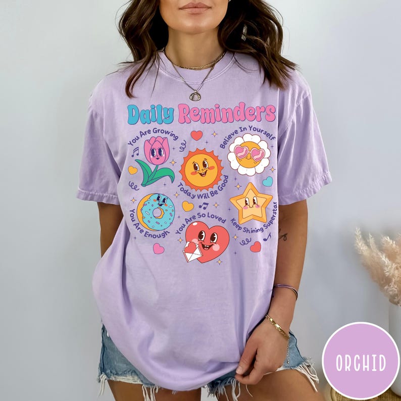 May include: A light purple t-shirt with the words "Daily Reminders" in a colorful, playful font. The shirt features various cartoon graphics, including a flower, sun, star, donut, and heart, each with an encouraging message.