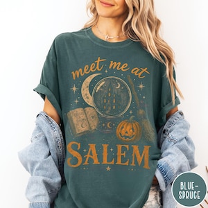 May include: A dark teal short-sleeve t-shirt featuring a graphic print in orange and cream. The design includes a crystal ball with a house inside, a crescent moon, an open book labeled Spells & Charms, a pumpkin, and a broom, with the text meet me at SALEM.
