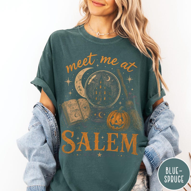 May include: A dark teal short-sleeve t-shirt featuring a graphic print in orange and cream. The design includes a crystal ball with a house inside, a crescent moon, an open book labeled Spells & Charms, a pumpkin, and a broom, with the text meet me at SALEM.