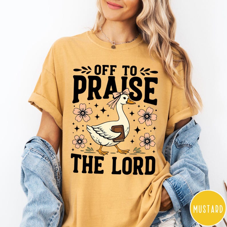 Puede incluir: Camiseta de color mostaza con el texto "OFF TO PRAISE THE LORD". El gr&aacute;fico presenta un ganso blanco sosteniendo un libro, adornado con un lazo rosa, rodeado de flores rosas y estrellas. La camiseta tiene las mangas enrolladas.