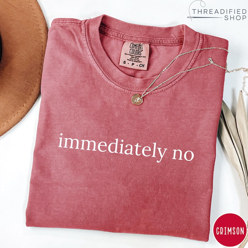 May include: Crimson-colored t-shirt with the phrase "immediately no" in white. The shirt has a crew neck and a Comfort Colors tag. A silver necklace with a pendant is draped across the shirt.