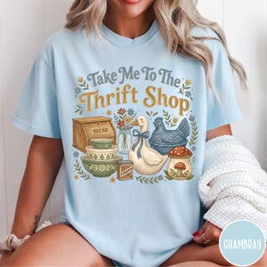 Op de afbeelding: Lichtblauw T-shirt met de tekst "Take Me To The Thrift Shop" in een decoratief lettertype. Het shirt heeft illustraties van een brooddoos, kommen, een eend, een kip, een paddenstoel en een pot bloemen.