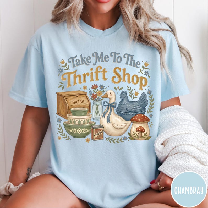 Op de afbeelding: Lichtblauw T-shirt met de tekst "Take Me To The Thrift Shop" in een decoratief lettertype. Het shirt heeft illustraties van een brooddoos, kommen, een eend, een kip, een paddenstoel en een pot bloemen.