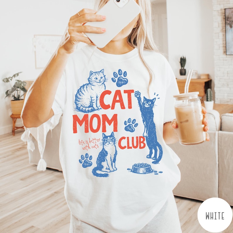 K&ouml;nnte beinhalten: Wei&szlig;es T-Shirt mit einem blau-roten Grafikdesign mit dem Text "CAT MOM CLUB" und Illustrationen von Katzen und Pfotenabdr&uuml;cken. Der Satz "life is better with cats" ist ebenfalls enthalten.
