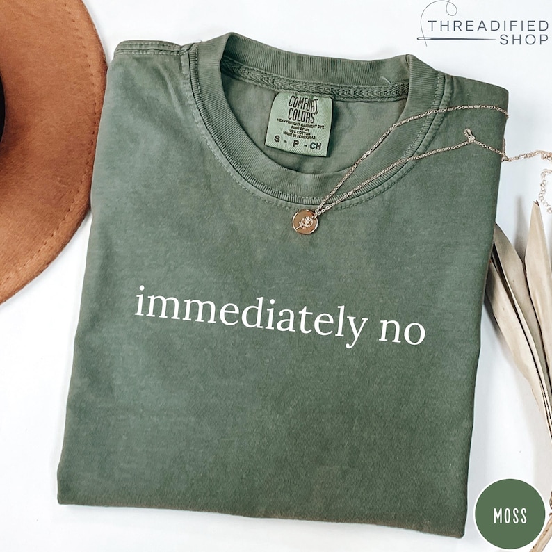 May include: A moss green t-shirt with the words "immediately no" printed in white. The shirt has a crew neck and short sleeves. A gold necklace with a pendant rests on the shirt. The shirt is folded.