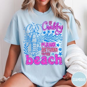 Puede incluir: Camiseta azul claro con un dise&ntilde;o gr&aacute;fico que presenta una langosta, olas y el texto "If Crabby Please Return to the Beach" en rosa y azul. El dise&ntilde;o tiene un aspecto vintage.