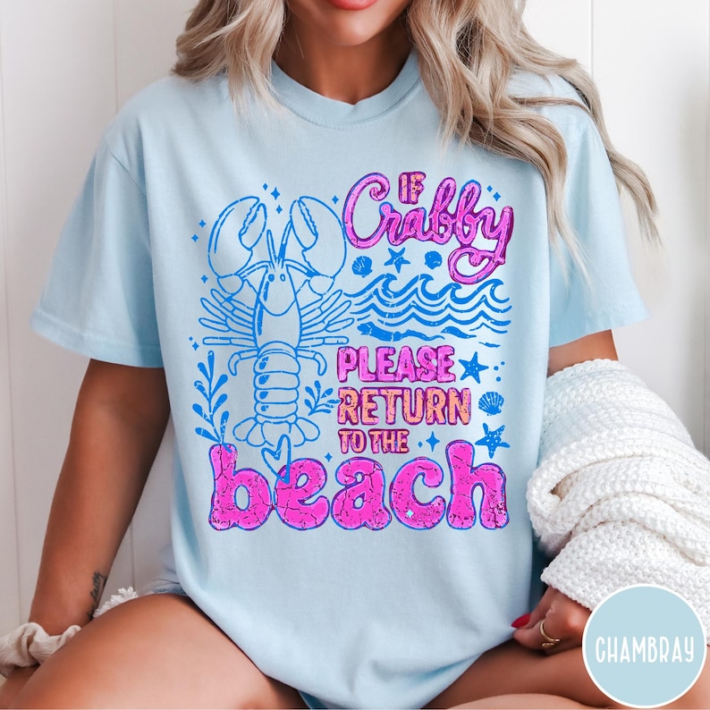Puede incluir: Camiseta azul claro con un dise&ntilde;o gr&aacute;fico que presenta una langosta, olas y el texto "If Crabby Please Return to the Beach" en rosa y azul. El dise&ntilde;o tiene un aspecto vintage.