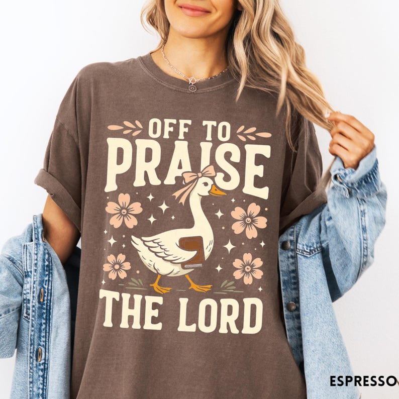 Puede incluir: Camiseta marr&oacute;n con el texto "OFF TO PRAISE THE LORD". El dise&ntilde;o presenta un ganso con lazo, flores y estrellas. La camiseta es de estilo casual.
