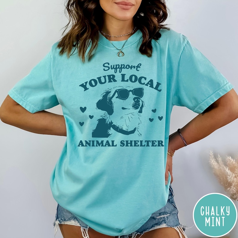 May include: A turquoise blue t-shirt with the text "Support Your Local Animal Shelter" in dark blue. A cartoon dog wearing sunglasses is in the center of the text. There are small hearts around the dog.