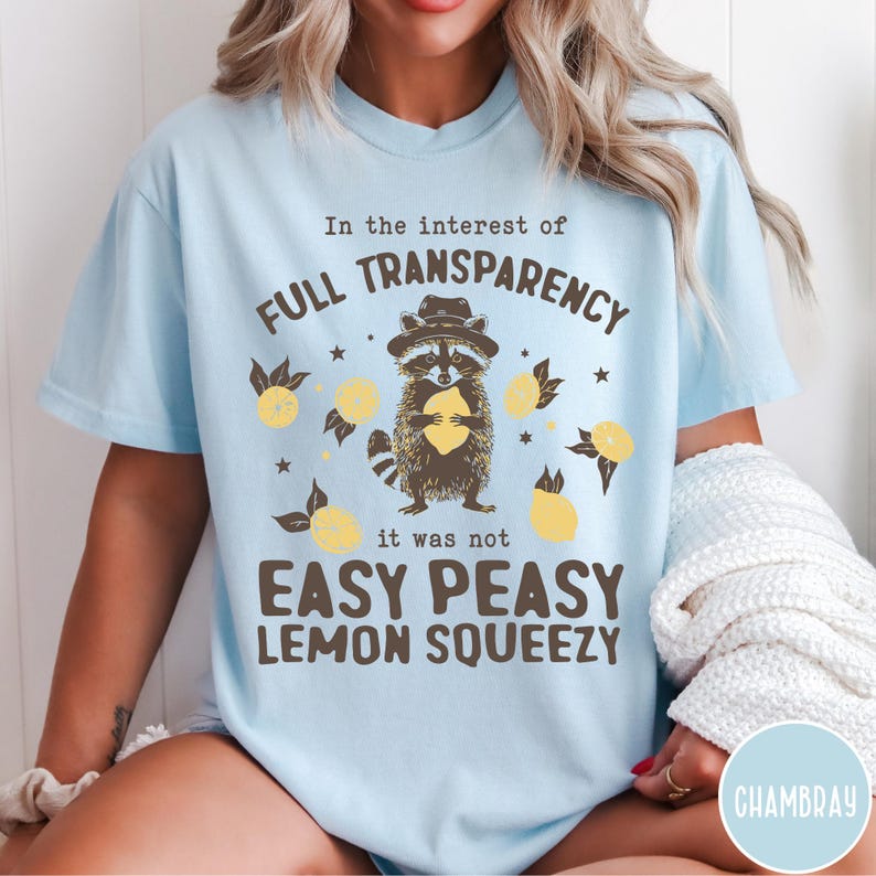 May include: Light blue t-shirt with a raccoon graphic holding a lemon. The text on the shirt reads "In the interest of full transparency it was not easy peasy lemon squeezy".