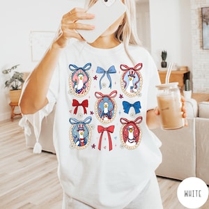 May include: White t-shirt featuring a repeating pattern of cartoon geese in patriotic outfits, framed by decorative bows. The geese are depicted with sunglasses, hats, and holding American flags, in a playful and festive design. The shirt is a comfortable, casual style.