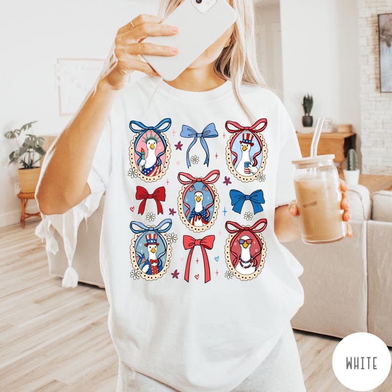 May include: White t-shirt featuring a repeating pattern of cartoon geese in patriotic outfits, framed by decorative bows. The geese are depicted with sunglasses, hats, and holding American flags, in a playful and festive design. The shirt is a comfortable, casual style.