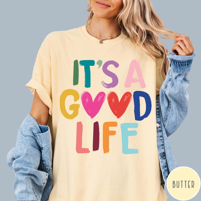 K&ouml;nnte beinhalten: Ein hellgelbes Kurzarm-T-Shirt mit dem mehrfarbigen Schriftzug IT'S A GOOD LIFE, bei dem zwei Herzen den Buchstaben O ersetzen. Das T-Shirt wird unter einer blauen Jeansjacke getragen und ein kleines kreisf&ouml;rmiges Etikett zeigt BUTTER.