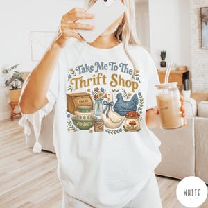 Op de afbeelding: Wit T-shirt met de tekst "Take Me To The Thrift Shop" en een vintage afbeelding. De afbeelding bevat een brooddoos, kommen, een eend, een kip en een paddenstoel. Het shirt heeft een casual stijl.