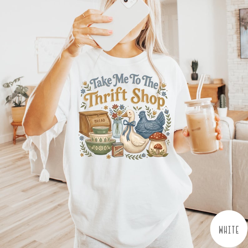 Op de afbeelding: Wit T-shirt met de tekst "Take Me To The Thrift Shop" en een vintage afbeelding. De afbeelding bevat een brooddoos, kommen, een eend, een kip en een paddenstoel. Het shirt heeft een casual stijl.