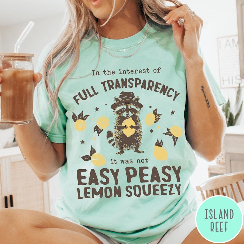 May include: A turquoise blue t-shirt with a raccoon graphic holding a lemon. The text on the shirt reads "In the interest of full transparency it was not easy peasy lemon squeezy".