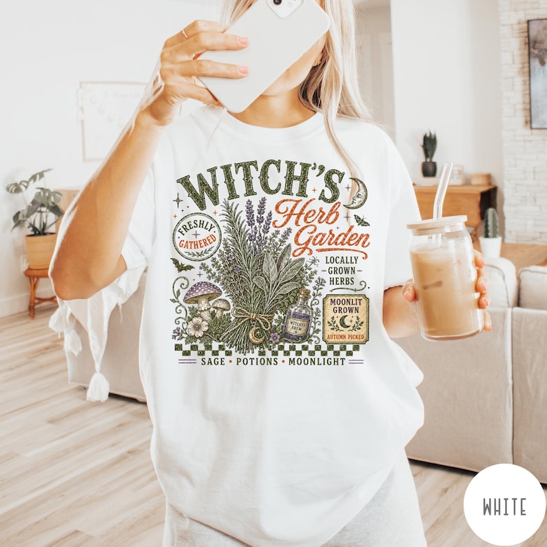 May include: A white t-shirt featuring a graphic print with the text Witch's Herb Garden. The design includes illustrations of a bundle of herbs, a mushroom, a small bottle, a crescent moon, and text reading Freshly Gathered, Locally Grown Herbs, and Moonlit Grown.