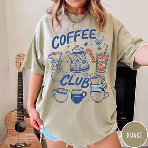 May include: Khaki-colored t-shirt with a "Coffee Club" graphic. The design features a blue teapot, coffee cups, and a glass of iced coffee. The text "all day, every day" is also included in the design.
