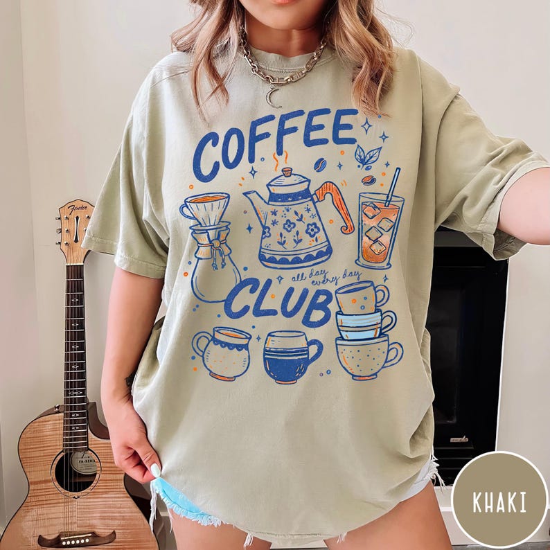 May include: Khaki-colored t-shirt with a "Coffee Club" graphic. The design features a blue teapot, coffee cups, and a glass of iced coffee. The text "all day, every day" is also included in the design.