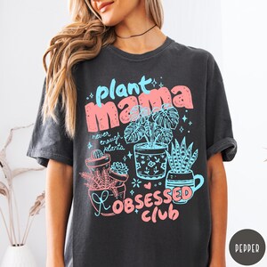 Puede incluir: Camiseta gris oscuro con un gr&aacute;fico colorido. El dise&ntilde;o presenta las palabras "plant mama" en rosa y azul, junto con ilustraciones de plantas en macetas y el texto "Obsessed Club". Una camiseta divertida e informal para los amantes de las plantas.