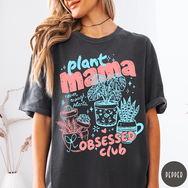 Puede incluir: Camiseta gris oscuro con un gr&aacute;fico colorido. El dise&ntilde;o presenta las palabras "plant mama" en rosa y azul, junto con ilustraciones de plantas en macetas y el texto "Obsessed Club". Una camiseta divertida e informal para los amantes de las plantas.