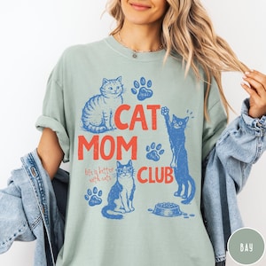 K&ouml;nnte beinhalten: Hellgr&uuml;nes T-Shirt mit dem Schriftzug "CAT MOM CLUB" in Rot und Blau. Das Design zeigt Illustrationen von Katzen, Pfotenabdr&uuml;cken und den Worten "life is better with cats".