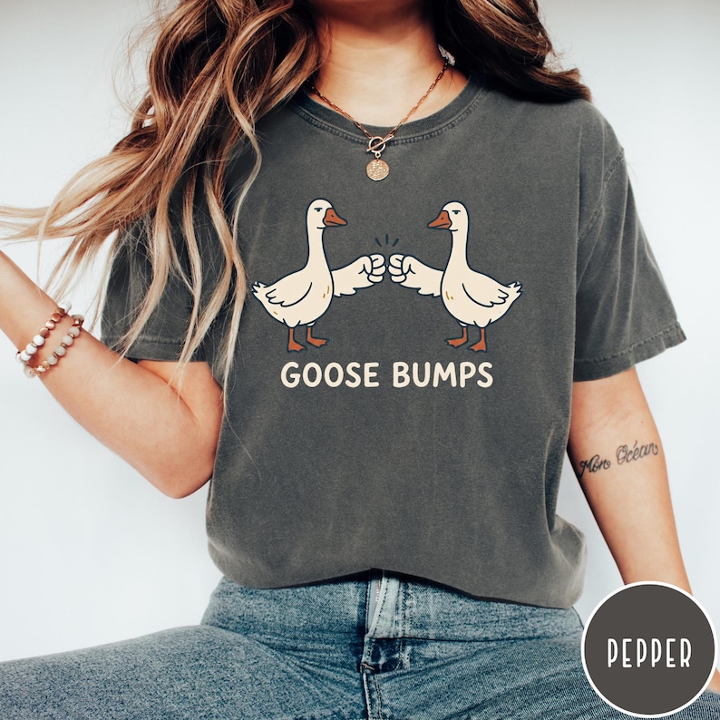 Puede incluir: Una camiseta de manga corta en color gris carb&oacute;n con un gr&aacute;fico de dos gansos blancos chocando los pu&ntilde;os. Debajo de los gansos, el texto GOOSE BUMPS est&aacute; impreso en una fuente negrita de color crema. La camiseta tiene cuello redondo.