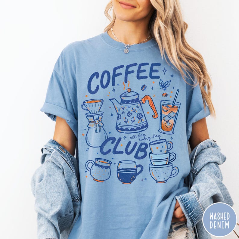 May include: Light blue t-shirt with a coffee-themed graphic. The design includes a teapot, coffee cups, and the words "COFFEE CLUB" and "all day, every day." The shirt has a washed denim look.