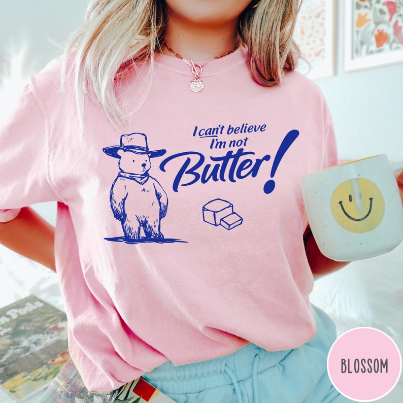 May include: Pink t-shirt with a blue graphic of a bear wearing a cowboy hat and the text "I can't believe I'm not Butter!"
