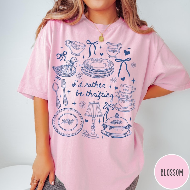 May include: A pale pink t-shirt featuring a blue illustration of vintage crockery, a duck, and the words "I'd rather be thrifting." The shirt has a round neck and short sleeves. The word "Blossom" is in a pink circle.