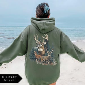Op de afbeelding: Militairgroene hoodie met een hertenafbeelding op de rug. De afbeelding toont een bok met grote geweien en twee kleinere herten in een bos. De hoodie heeft een capuchon en lange mouwen. De tekst "MILITARY GREEN" staat onderaan.