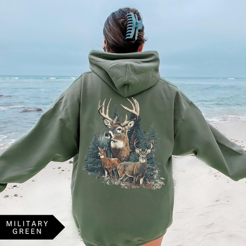 Op de afbeelding: Militairgroene hoodie met een hertenafbeelding op de rug. De afbeelding toont een bok met grote geweien en twee kleinere herten in een bos. De hoodie heeft een capuchon en lange mouwen. De tekst "MILITARY GREEN" staat onderaan.