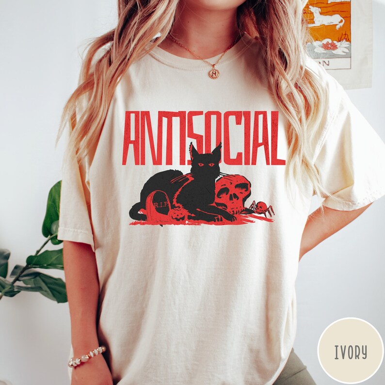 Może przedstawiać: Biały T-shirt z napisem "ANTISOCIAL" w czerwonych literach. Czarny kot z czerwonymi oczami siedzi na czaszce z pająkiem obok. Napis "R.I.P." znajduje się na nagrobku za kotem.
