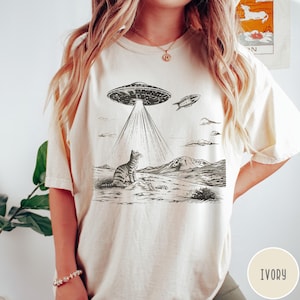 Pode incluir: Uma camiseta branca com uma ilustra&ccedil;&atilde;o em preto e branco de um gato olhando para cima para um OVNI. O OVNI est&aacute; emitindo raios de luz. O texto "IVORY" est&aacute; no canto inferior direito da imagem.