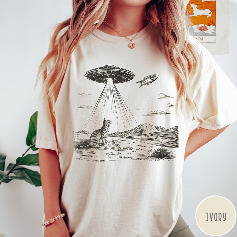 Pode incluir: Uma camiseta branca com uma ilustra&ccedil;&atilde;o em preto e branco de um gato olhando para cima para um OVNI. O OVNI est&aacute; emitindo raios de luz. O texto "IVORY" est&aacute; no canto inferior direito da imagem.