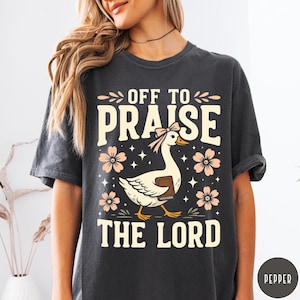 Puede incluir: Camiseta gris oscuro con el texto "OFF TO PRAISE THE LORD". El dise&ntilde;o incluye un ganso blanco con un lazo rosa y un libro, rodeado de flores rosas y estrellas. Una camiseta casual y c&oacute;moda.