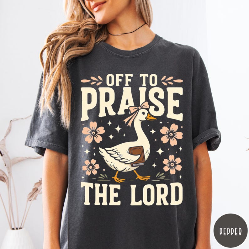 Puede incluir: Camiseta gris oscuro con el texto "OFF TO PRAISE THE LORD". El dise&ntilde;o incluye un ganso blanco con un lazo rosa y un libro, rodeado de flores rosas y estrellas. Una camiseta casual y c&oacute;moda.