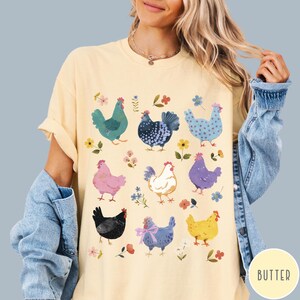 K&ouml;nnte beinhalten: Ein gelbes T-Shirt mit einem bunten Muster aus H&uuml;hnern und Blumen. Die H&uuml;hner sind in verschiedenen Farben und Posen dargestellt, darunter stehend, gehend und sitzend. Die Blumen sind in Rosa-, Lila- und Blaut&ouml;nen gehalten.