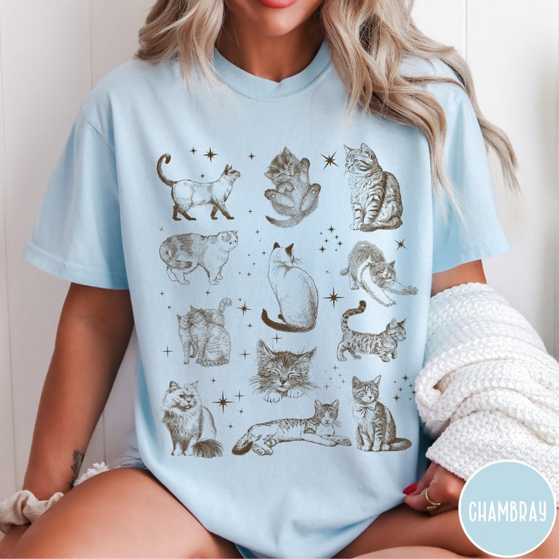 K&ouml;nnte beinhalten: Ein hellblaues T-Shirt mit einem Aufdruck von verschiedenen Katzen in verschiedenen Posen. Die Katzen sind im Vintage-Stil gezeichnet und von Sternen umgeben.